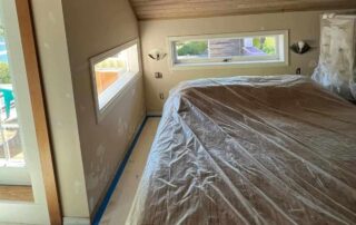 Taupe-painted residential bedroom with wood ceiling, horizontal windows, white trim, and protective plastic sheeting on floor